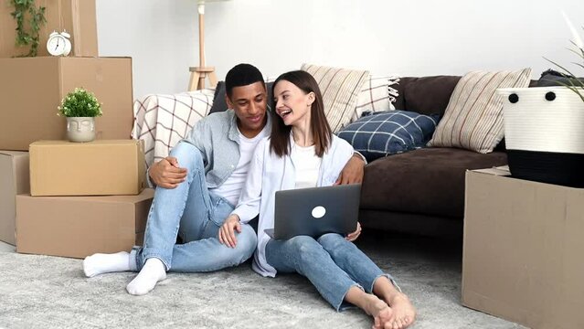 Happy joyful multiracial married couple in love, bought new housing, sit among boxes with things for home, use laptop, talk on video call with friends, show their new home, smiling