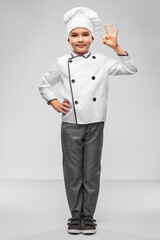 cooking, culinary and profession concept - happy smiling little boy in chef's toque and jacket showing ok gesture over grey background