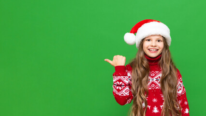 A child in a Christmas hat. A little girl in a Santa Claus hat and a festive sweater points to your advertisement on a green isolated background.