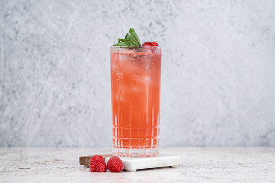 Bebida de frutos rojos con hielo y romero sobre fondo claro de textura de concreto, en vaso higball con decorado de frambuesa y menta, vista frontal