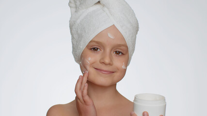 Lovely young child girl with towel on head applying cleansing moisturizing cream from creme jar....