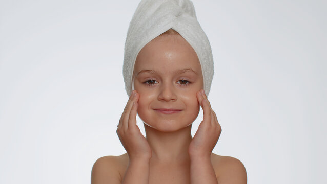 Lovely young child girl after bath in towel on head applying cleansing moisturizing cream. Teenager kid face skincare healthy treatment, natural creme cosmetics. Female portrait. Perfect fresh clean