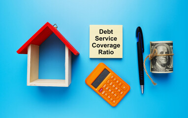 A picture house miniature, with fake money, Debt Service Coverage Ratio on notepad, calculator, and pen on blue background.