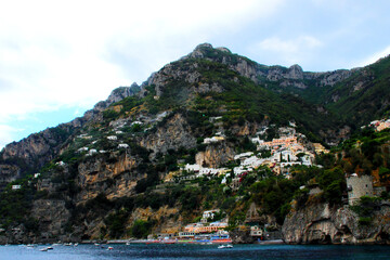 Fototapeta na wymiar Varied panorama in Amalfi coast with the cusp of a mountain crest high in the sky, a lot of mostly white buildings below, a stone tower on the right and a colorful waterfront near the boat-filled sea