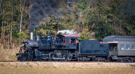 Fototapeta na wymiar A View of a Restored Steam Passenger Train Blowing Smoke and Steam Traveling Along a Rural Countryside on a Sunny Day