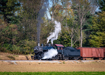 Fototapeta na wymiar A View of a Restored Steam Freight Train Blowing Smoke and Steam Traveling Along a Rural Countryside on a Sunny Day