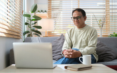 Obraz premium Retired man resting on couch in bright living room and reading online news on laptop at the morning.