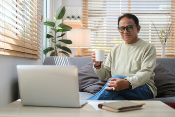 Positive mature man drinking coffee and using laptop while relaxing on couch in cozy home.