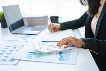 Happy Asian business woman using calculator, sitting at a desk in office with business chart paperwork.