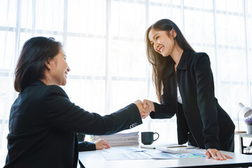 Two businesswoman shaking hands to congratulate success with graph document calculator placed at the office desk.
