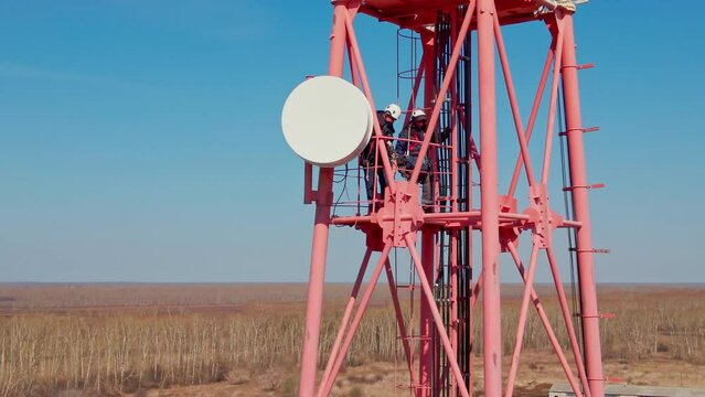 Two workers in helmet and overalls maintain and diagnose satellite dishes on metal tower. Dangerous work at altitude. Shooting from drone.