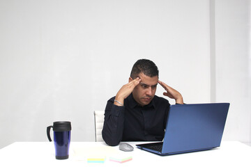 Dark-haired latin adult man works in his office with laptop with a lot of stress having a headache with a migraine and suffering from tiredness and burnout
