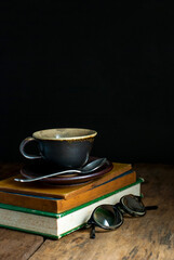 Coffee cup with saucer and coffee spoon placed on a pile of old books with glasses on a rustic...