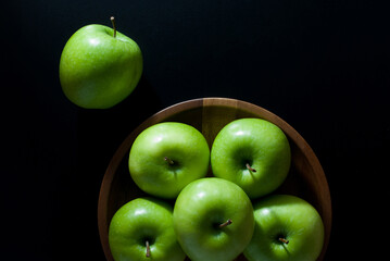 Green apples are placed in a wooden round tray and rest on a black background, top view.