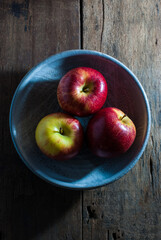 Three red apples in a ceramic bowl on a rustic wooden floor, top view.