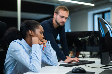 Colleagues look at the monitor and decide working moments. Caucasian man helps sad african woman solve computer problem.