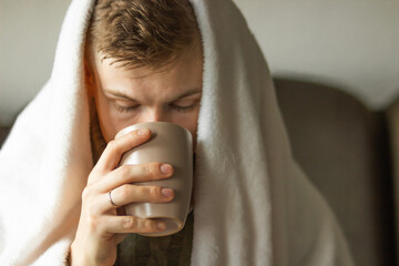 Caucasian young man has fever and common cold sitting at home, wrapped in warm blanket and drinking...