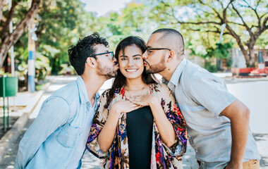 Two men kissing a girl cheek. Portrait of two guys kissing a girl cheek. Two young men kissing a woman cheek outdoor, love triangle concept. Polygamy concept - obrazy, fototapety, plakaty