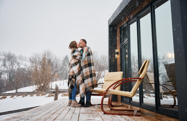 Man and woman wrapped in blanket kissing outdoors under winter snow. Happy couple sharing romantic moment while standing near chairs outside scandinavian house barnhouse with panoramic windows.