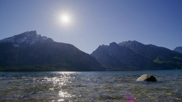 Crystal clear alpine lake with sunbeams reflecting on water surface. Craggy, snow-capped peaks of Teton Mountain Range with sunrise skyline in the background. High quality 4k footage