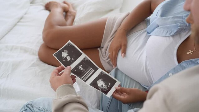 Multiracial pregnant couple watching ultrasound image picture at home. Closeup feeling happy, stroking tummy and excited on seeing baby ultrasound