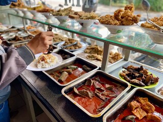 Padang food stalls share a variety of buffet menus with vegetables, chicken curry sauce, beef, eggs, tofu, tempeh. the waiter prepares the food. Menu in glass display case. Asia and asian food. Pile.