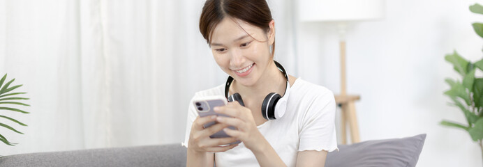 Beautiful Asian women are playing games on their mobile phones or smartphones with pleasure and fun, Put on headphones to play games, Girl's favorite corner on weekends,  Happy living room.