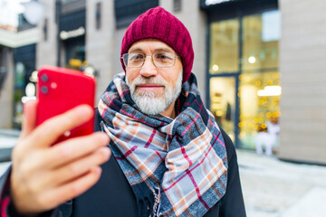 good looking mature man in hat and glasses using mobile phone app texting outside