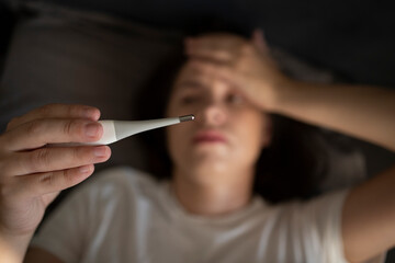 Woman is lying in bed and holding her head in her hands and sees an increased temperature on the thermometer. Concept: deprivation, flu, cold, coronovirus, flu epidemic. 