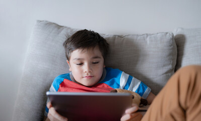 School kid using tablet readying story from internet, Happy boy sitting on sofa doing studying online learning  at home, Child holding digital pad using wifi playing game or chatting with friends.