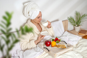 Obraz na płótnie Canvas Woman eating fruit breakfast in bed at home in the morning. Lady wrapping a towel around head. Girl wearing bathrobe on clean white bedding with cozy blanket. Healthy lifestyle