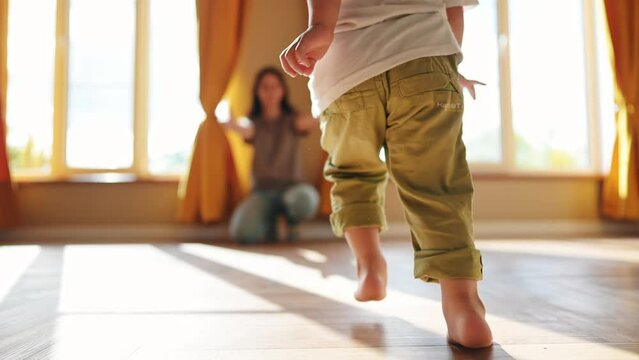 baby runs to mom at home. a child with bare legs runs across the floor to his mother against a sunny window. happy family kid dream concept. kid running back view at lifestyle home hugging mom