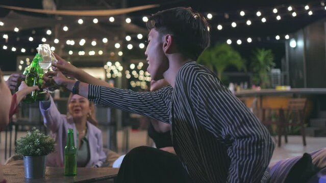 Happy LGBTQ and Asian friends clinking alcohol bottles celebrating at night club rooftop