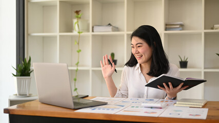 Cheerful asian female entrepreneur making video call with her business partner or watching online webinar on laptop computer
