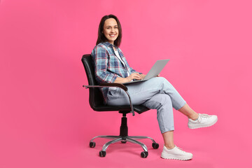 Mature woman with laptop sitting in comfortable office chair on pink background