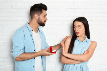 Young woman rejecting engagement ring from boyfriend near white brick wall