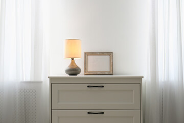 Modern chest of drawers with lamp near window in room