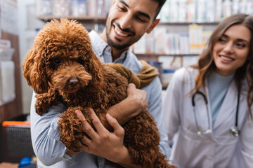 Cheerful arabian man looking at poodle near veterinarian in pet shop