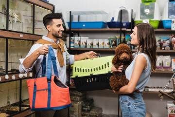 Side view of cheerful interracial couple holding bag and poodle in pet shop