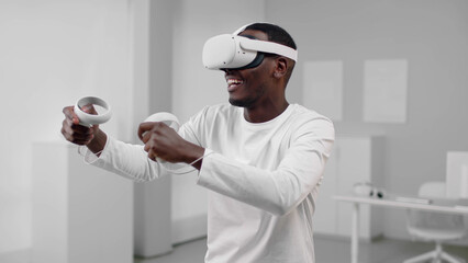African-American man in white clothes play computer game in vr headset using joysticks