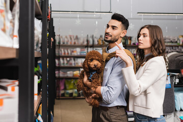 Woman pointing with finger near muslim boyfriend holding poodle in pet shop