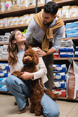 Positive multiethnic couple with brown poodle looking at each other in pet shop