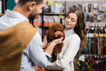Smiling woman holding poodle near blurred arabian boyfriend in pet shop