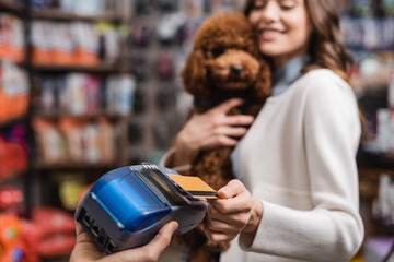 Blurred woman holding poodle and paying with credit card in pet shop