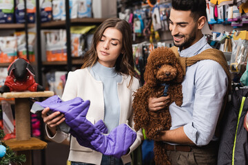 Smiling muslim man holding poodle near girlfriend with animal jacket in shop
