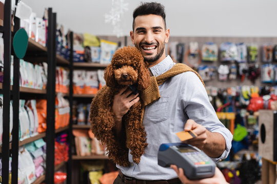 Positive arabian man holding poodle and looking at camera while paying with credit card in pet shop