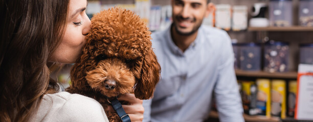 Woman kissing brown poodle near blurred seller in pet shop, banner