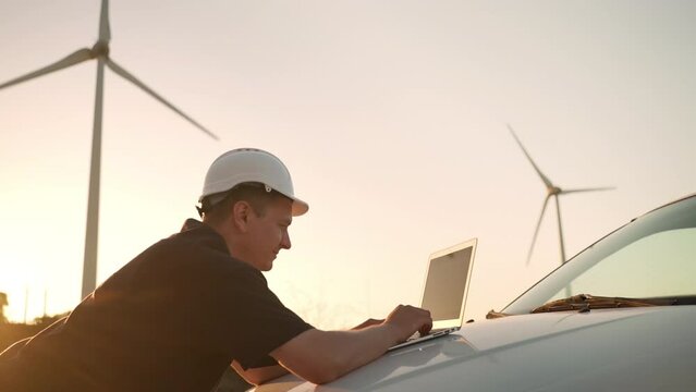 Technician engineer working at wind turbine using laptop computer worker or operator checks wind turbine farm does maintenance to power generator station lean on car wind turbine and sunset background