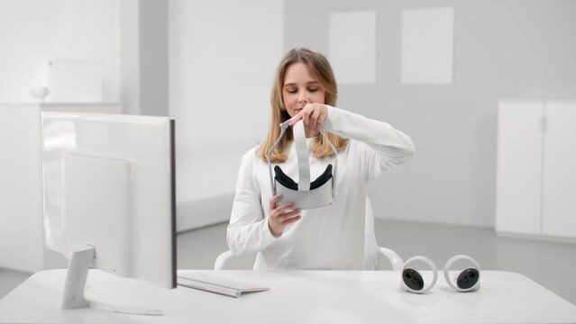 Young woman in white clothes put on vr headset sitting at desk in white room