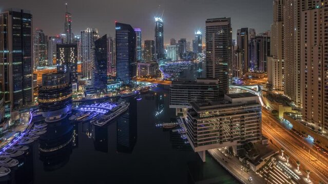 Aerial panoramic view to Dubai marina illuminated skyscrapers around canal with floating yachts timelapse during all night and lights turning off. White boats are parked in yacht club